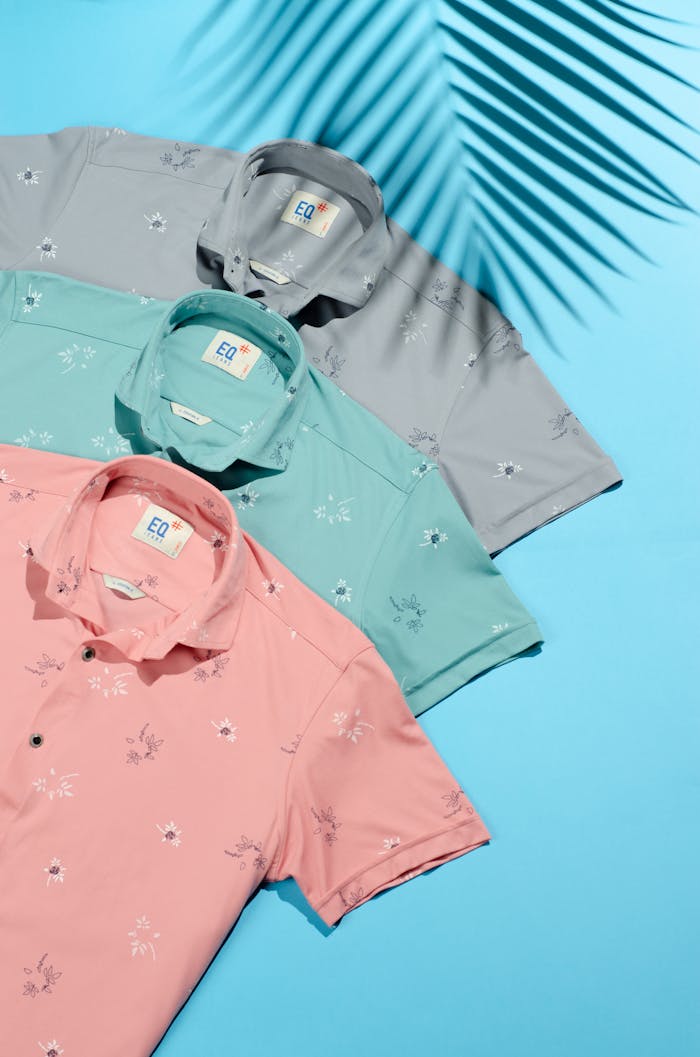 Three stylish polo shirts on a blue background with tropical shadow play.